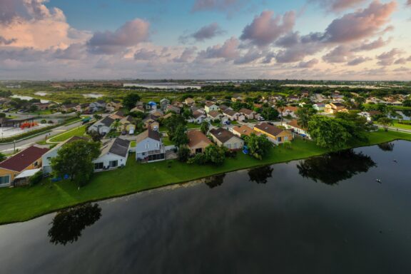 Aerial view of a residential area in Miramar, Florida, with houses, streets, and greenery, adjacent to a body of water, under a cloudy sky at dusk.