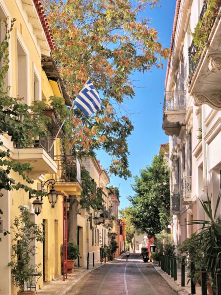 A narrow street in Athens, Greece, lined with colorful buildings and a Greek flag hanging from a balcony, with trees and a clear sky above.