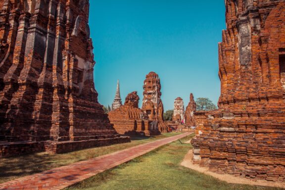 A photo of ancient ruins in Ayutthaya, Thailand, featuring weathered brick structures and pathways under a clear blue sky.