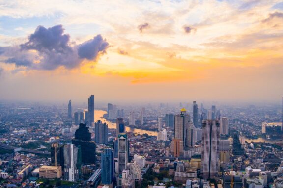 Aerial view of Bangkok city skyline at dusk with skyscrapers and a river winding through the city, under a sky with clouds tinged by the setting sun.