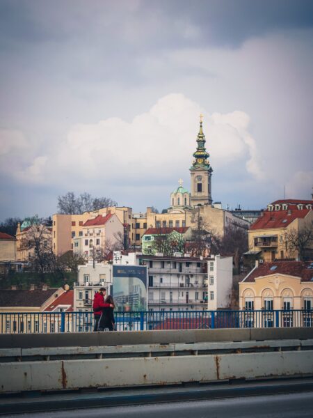 A person walking across a bridge with a view of Belgrade, Serbia's cityscape in the background, featuring historical buildings and a church spire.