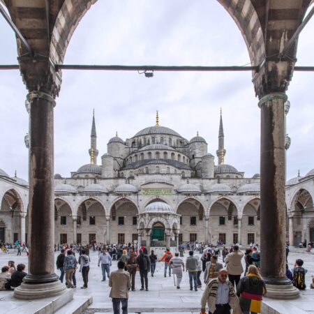 A view of the Blue Mosque in Turkey, seen through arches with visitors walking in the courtyard.