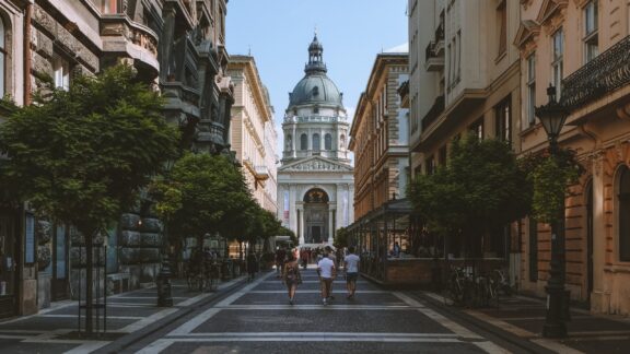 A street view in Budapest, Hungary, with people walking, buildings on either side, and a large domed structure in the background.