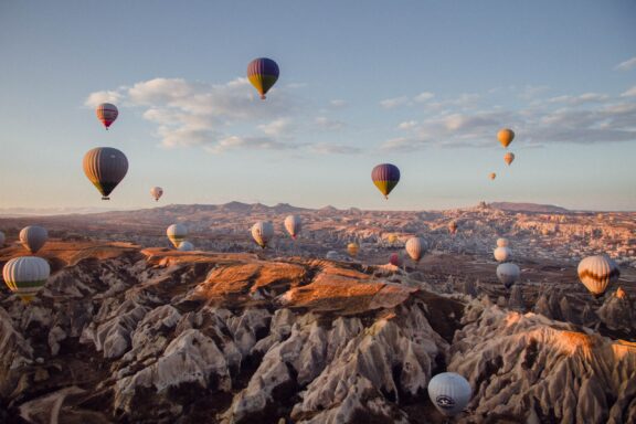 A scenic view of numerous hot air balloons floating above the rocky landscape of Cappadocia, Turkey, during sunrise or sunset.