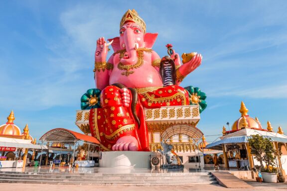 A vibrant statue of Hindu deity Ganesha sits in lotus position in Chachoengsao, Thailand, with traditional Thai buildings behind.