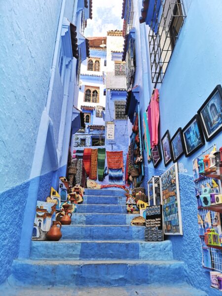 A narrow stairway in Chefchaouen, Morocco, with blue walls, lined with colorful textiles, pottery, and paintings for sale.