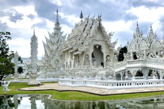 A white ornate temple with intricate architectural details in Chiang Rai, Thailand, reflected in a nearby water body under a partly cloudy sky.