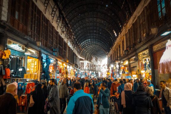 A bustling market scene with numerous people walking and shopping under a covered walkway in Damascus, Syria.