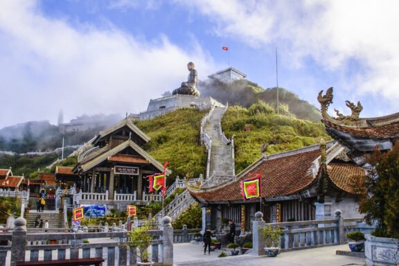 A picturesque view of Fansipan, Vietnam, showcasing traditional architecture, a large statue, and the national flag on a cloudy hilltop.