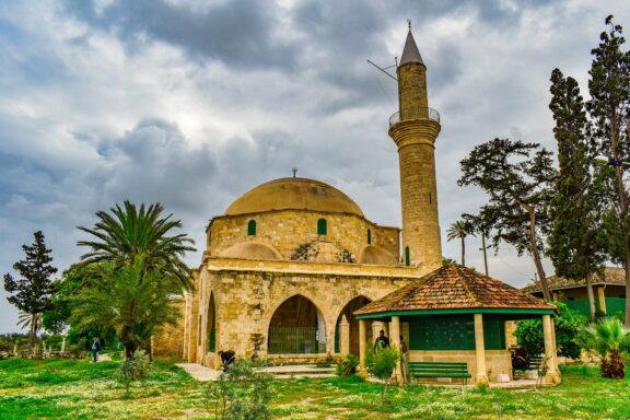 A mosque with a single minaret surrounded by greenery under a cloudy sky, located at Hala Sultan Tekke.