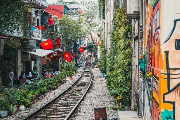 A narrow Hanoi street has train tracks, buildings with Vietnamese flags, and a graffiti-covered wall.