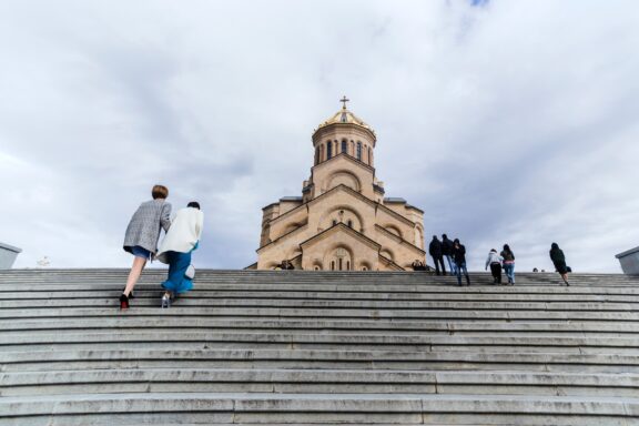 A couple ascends the steps of Tbilisi's Holy Trinity Cathedral, Georgia, amidst scattered visitors and cloudy skies.