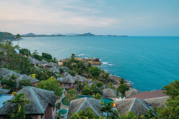 A coastal scene in Koh Samui, Thailand, with thatched bungalows, a calm blue sea at dusk, and hills in the background.