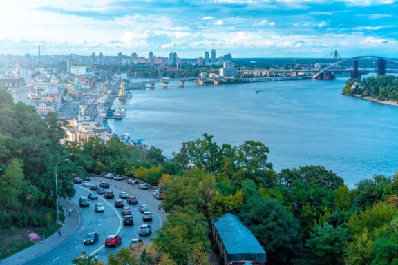 Aerial view of a cityscape with a river, bridges, and buildings, with a winding road in the foreground, likely in Kyiv, Ukraine.