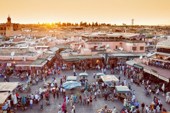 Sunset over a bustling market square in Marrakesh, Morocco, with crowds of people, stalls, and buildings under a warm sky.