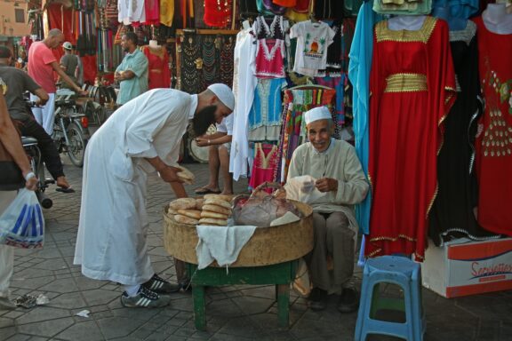 Two men in traditional Moroccan attire are interacting at a street market, one handling bread, the other sitting nearby.