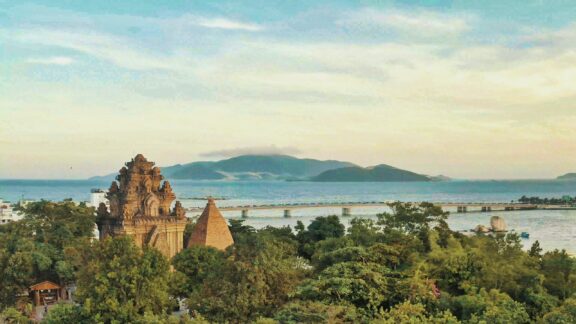 A picturesque view of Nha Trang, Vietnam, showcasing a large temple, calm sea, mountains, and a sunset sky.