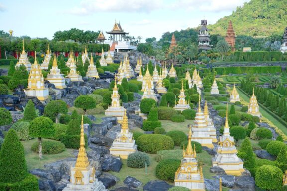 Nong Nooch Tropical Garden in Thailand features golden pagodas, green bushes, and traditional Thai architecture under a cloudy sky.