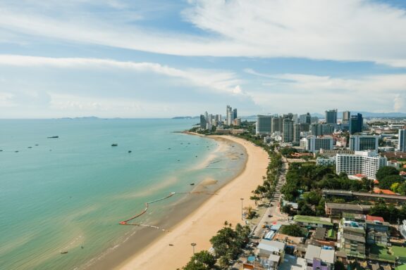 Aerial view of Pattaya beach and skyline with buildings along the coast, boats in the water, and a clear sky, in Thailand.