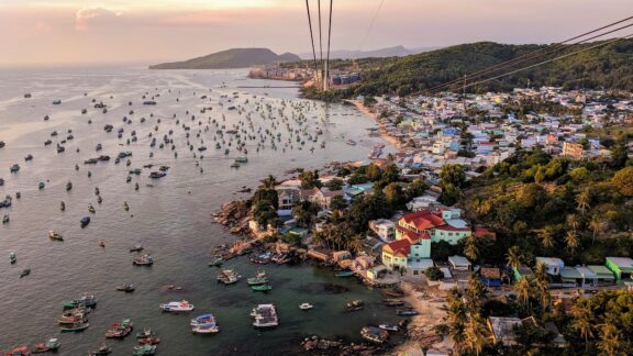 Aerial view of a coastal town in Phu Quoc, Vietnam, with numerous boats in the water and buildings nestled among greenery at dusk.