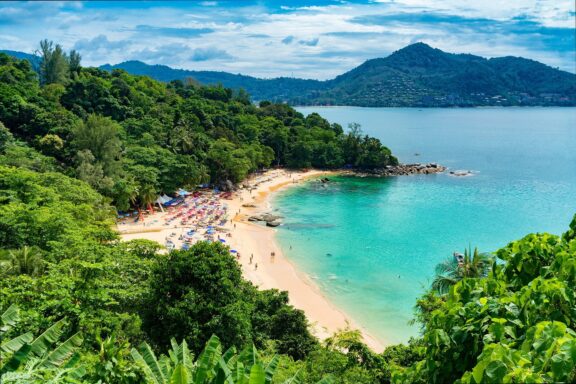 A scenic view of a beach in Phuket, Thailand, with turquoise waters, sandy shore, lush greenery, and visitors enjoying the sun.