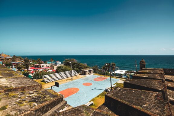 A vibrant basketball court by the sea in San Juan, Puerto Rico, with historical forts in the foreground and a clear blue sky.