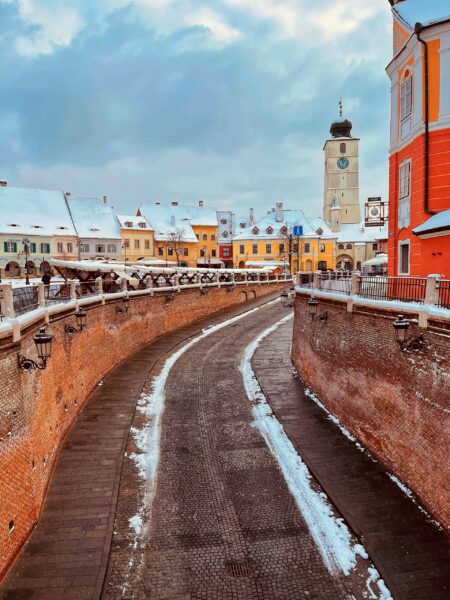 A cobblestone bridge with snow on the sides leading towards a colorful European town with historical buildings under a cloudy sky, in Sibiu, Romania.