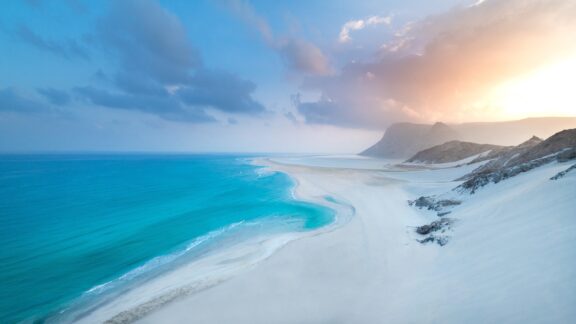 A panoramic view of a serene beach on Socotra Island, Yemen, with white sands and turquoise waters under a dramatic sky at sunset.