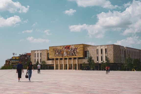 A group of people walking across a spacious square towards a large building with a mural in Tirana, Albania, under a partly cloudy sky.