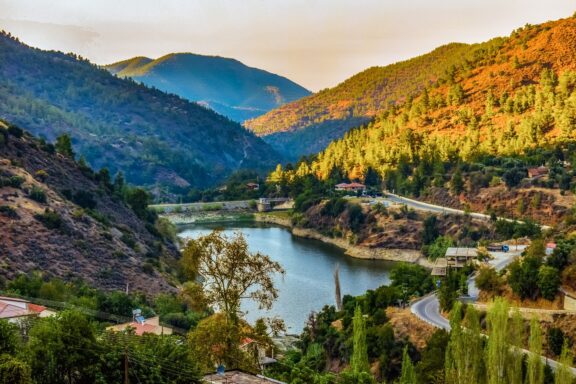 A scenic view of the Troodos Mountains in Cyprus, featuring a winding river, lush greenery, and mountainous terrain bathed in warm sunlight.