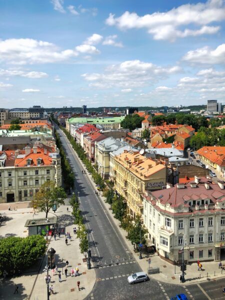 Aerial view of a tree-lined street in Vilnius, Lithuania, with historic buildings on either side under a partly cloudy sky.