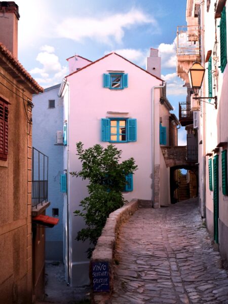 A cobblestone street in Vrbnik, Croatia features a pink building on the right and stone buildings on the left under a semi-cloudy sky.