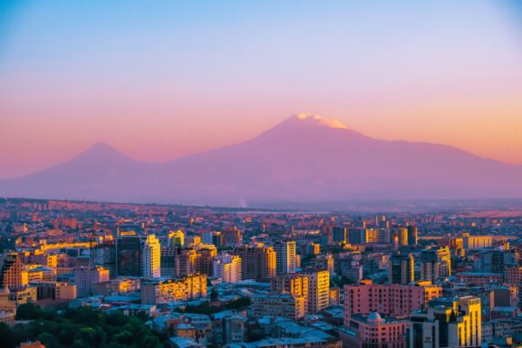 A panoramic view of Yerevan, Armenia during sunset with Mount Ararat in the background and the city illuminated by warm light.