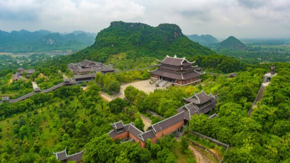 Aerial view of Bai Dinh Temple Spiritual and Cultural Complex in a lush, green landscape with mountains in the background, under a cloudy sky.