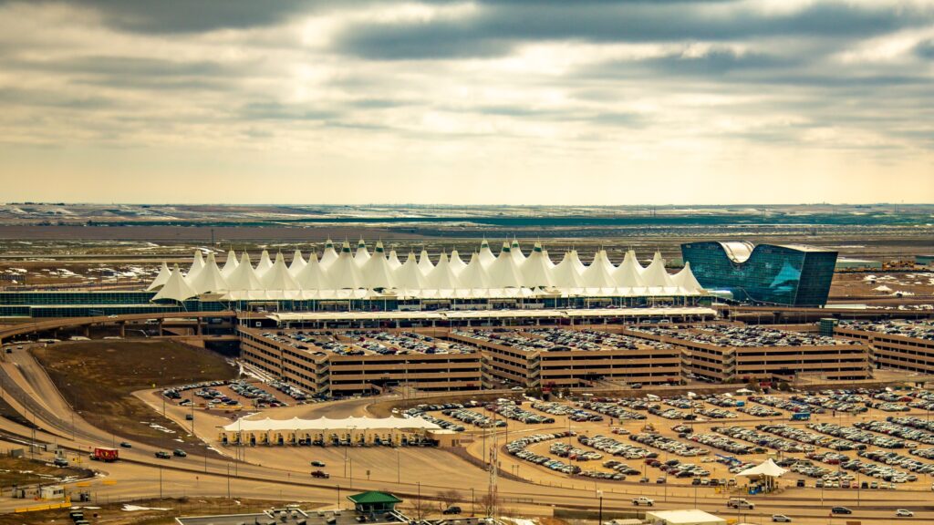 Aerial view of Denver Airport terminal with distinctive peaked roof design, surrounded by parking lots, under a cloudy sky.