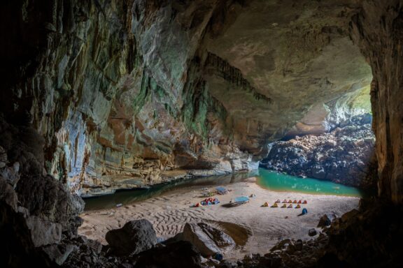 A large, naturally lit cave with a beach and water body, featuring kayaks and life vests, located in Doong Cave, Vietnam.