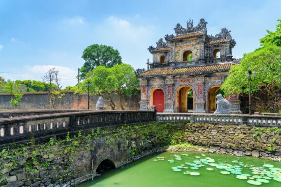 A traditional Vietnamese gate with intricate carvings, red doors, stone statues, and a lily pad-covered moat at Hien Nhon Gate, Vietnam.