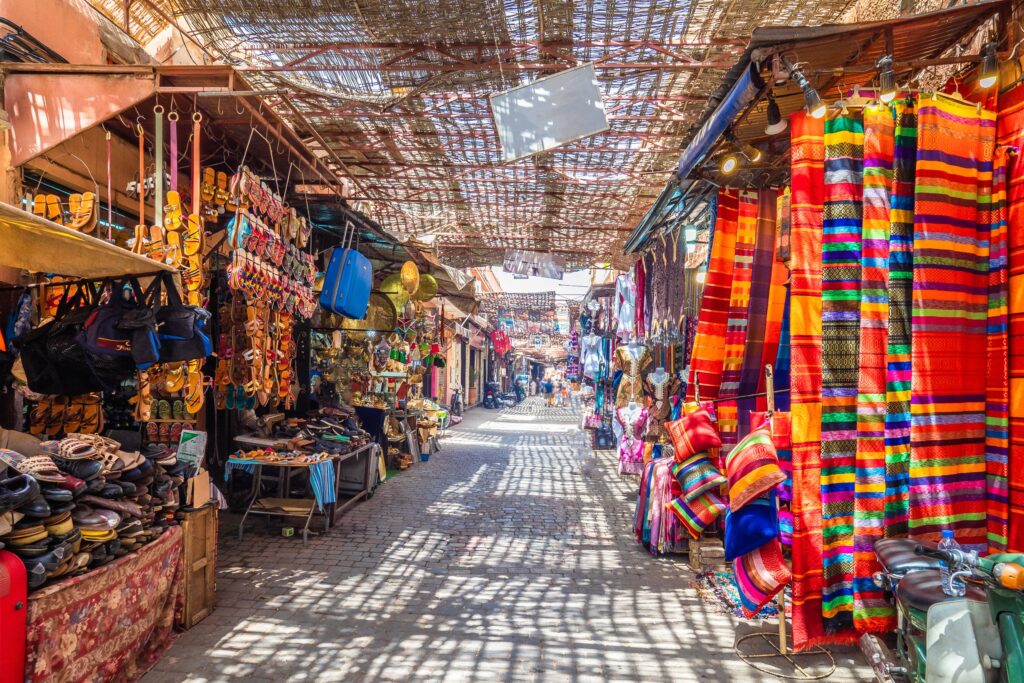 A vibrant market scene at Jamaa el Fna, Morocco, with colorful souvenirs displayed on either side under a shaded walkway.