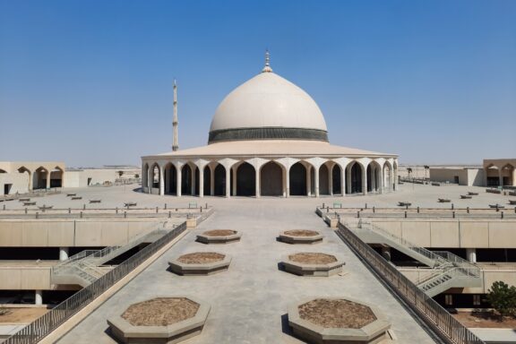 A mosque with a white dome and minaret near Dammam Airport, with a pathway and geometric planters in front.