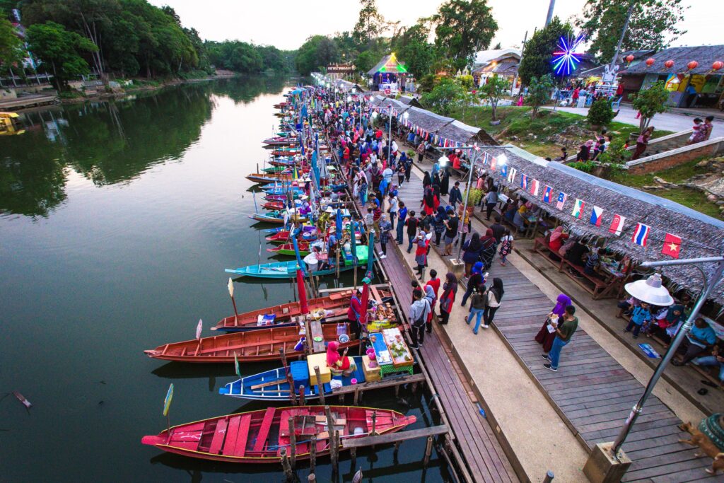 A vibrant evening market along a river in Songkhla, Thailand, with numerous colorful boats docked beside the walkway and people browsing the stalls.