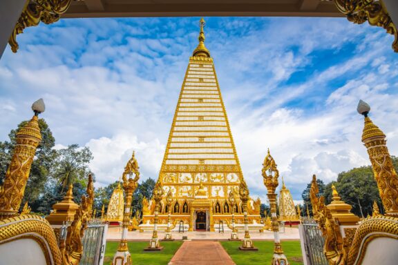 A golden and white striped pagoda at a temple in Ubon Ratchathani, Thailand, flanked by ornate golden sculptures under a blue sky with clouds.