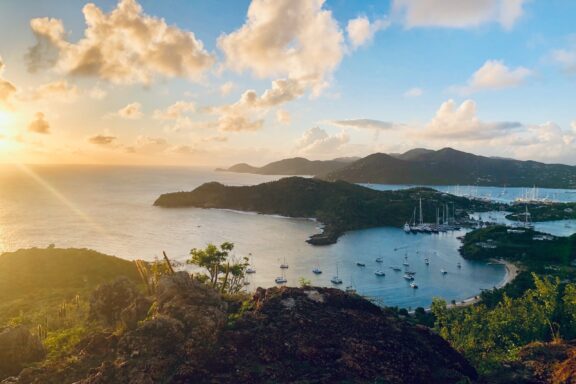 A picturesque view of Antigua and Barbuda's coastline, with boats in the bay, green hills, and a sunset-lit sky.