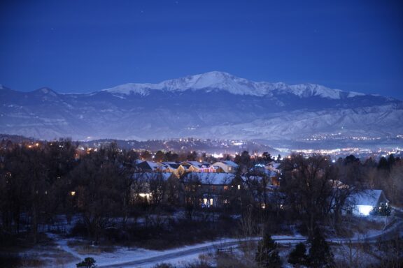 Twilight view of Colorado Springs with illuminated buildings in the foreground and the snow-capped Pikes Peak in the background.