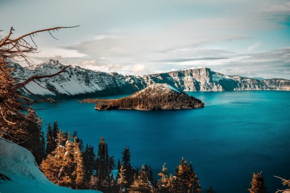 A peaceful scene at Oregon's Crater Lake, featuring a central island, steep cliffs, snowy slopes, and a semi-cloudy sky.