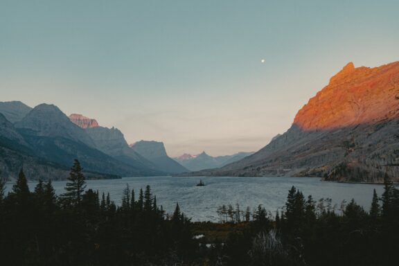 A serene dusk scene at Glacier National Park, Montana, featuring a calm lake with mountain silhouettes and a softly lit sky.