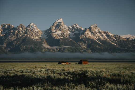 A peaceful scene at Wyoming's Grand Teton National Park with snow-capped mountains, fog, and wooden buildings in a grassy field.