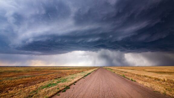 A vast, open landscape in Kansas with a dramatic storm cloud formation overhead, looming above a straight road that cuts through the scene.