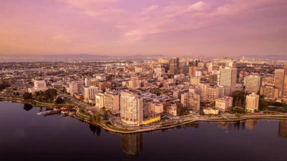 Aerial view of downtown Oakland, California at dusk with buildings reflecting the warm hues of the sunset and water in the foreground.