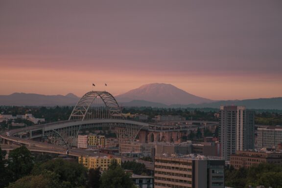 Sunset View of Portland, Oregon with Mount Hood in the Background A view of Portland, Oregon at dusk with a bridge in the foreground and Mount Hood in the background under a pastel-colored sky.