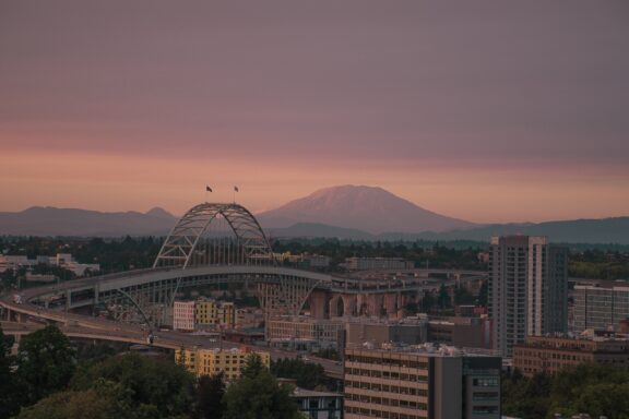 A view of Portland, Oregon at dusk with a bridge in the foreground and Mount Hood in the background under a pastel-colored sky.